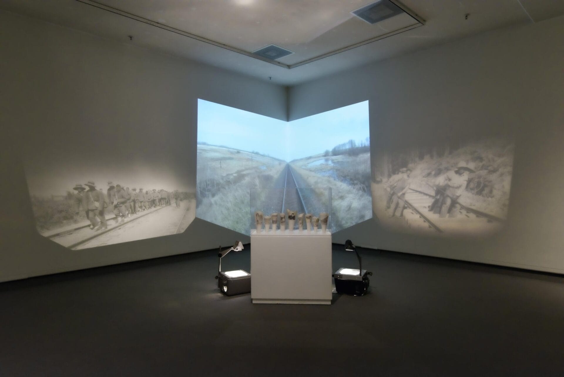 An art installation showcases a three-wall projection depicting scenes of railroads and workers. A central pedestal features small statues. Surrounding spotlights illuminate the display against the dimly-lit gallery backdrop.