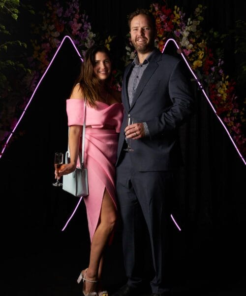 A man and woman stand together against a dark backdrop with vibrant flowers and pink neon lines forming a triangle shape. The woman wears a pink off-the-shoulder dress, and the man wears a dark suit. Both hold champagne glasses and smile at the camera.