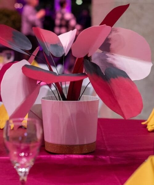A decorative table setup with a white pot containing large, pink and white artificial leaves. A wine bottle with a black and white label is placed beside the pot. The table is covered with a raspberry-colored cloth and is adorned with yellow napkins and glassware.