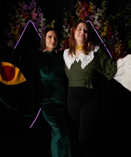 Two people stand in front of a backdrop adorned with colorful flowers and neon lights. The person on the left wears a green dress with a large, eye-shaped graphic on the sleeve, while the person on the right wears a green top with a white, ruffled collar and flared sleeves.