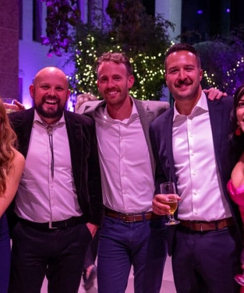 A group of five people dressed in formal attire stand together smiling at a lively evening event. Four individuals are holding drinks, while one person, second from the left, is not. Behind them, string lights and festive decorations are visible.