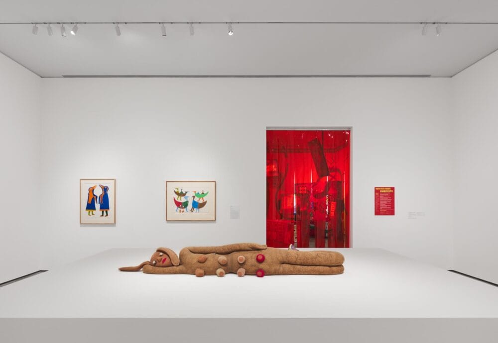 Une galerie d'art contemporain aux murs blancs expose une grande sculpture moelleuse représentant une figurine en pain d'épices allongée. Deux œuvres d'art encadrées et colorées sont accrochées au mur de gauche, et une installation éclairée en rouge est visible par une porte au fond.