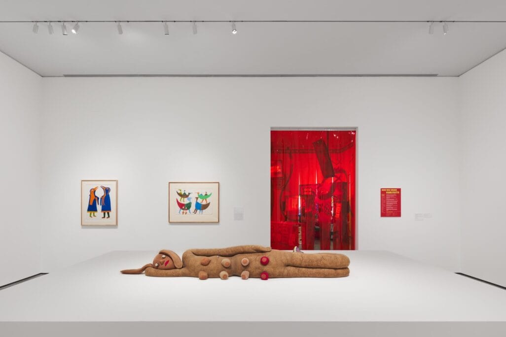 A large, whimsical sculpture resembling a gingerbread person lies on a white platform in an art gallery during RAWLCO Free Day, surrounded by colorful framed artworks and a red-lit room visible in the background.