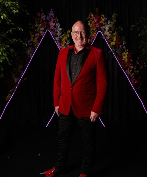A bald man wearing glasses stands smiling in front of a black background adorned with neon purple triangles and floral decorations. He is dressed in a black shirt, a red velvet blazer, black pants, and red shoes.