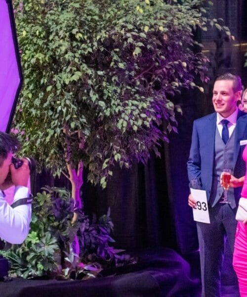 A man in a suit, holding a number card, and a woman in a bright pink dress pose for a photograph at a decorated event. The photographer is crouched down, taking their picture. The background features a large plant and decorative pink and neon lights.