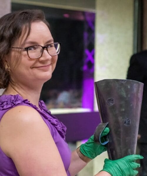 A woman in a sleeveless purple dress and green gloves smiles at the camera while holding a large dark metal object. Behind her, a man in a tuxedo speaks into a microphone at a podium. The background is blurred and features a mix of purple and neutral tones.