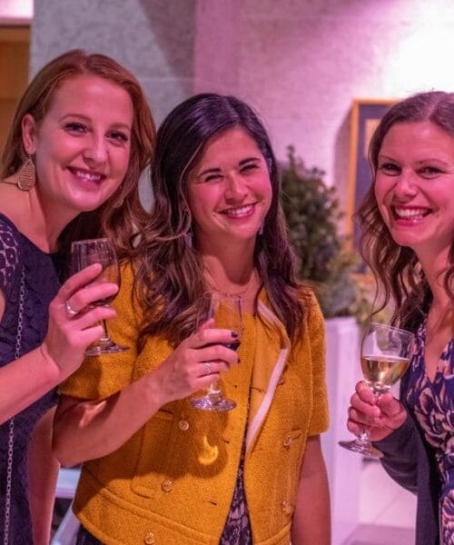 Three women are smiling and holding glasses of wine at a social event. All are dressed in casual yet elegant attire. The background is softly lit with purple lighting, giving a warm and inviting ambiance.