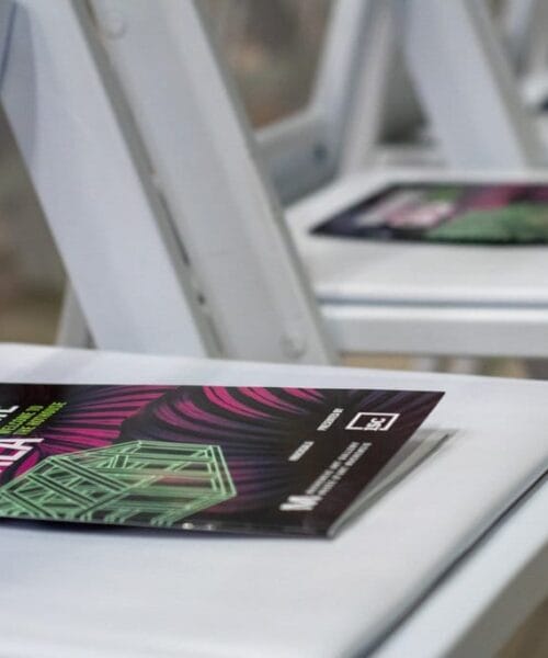 A row of white chairs, each with a folded event program placed on the seat. The program cover is predominantly black with a green building illustration and purple text that reads 