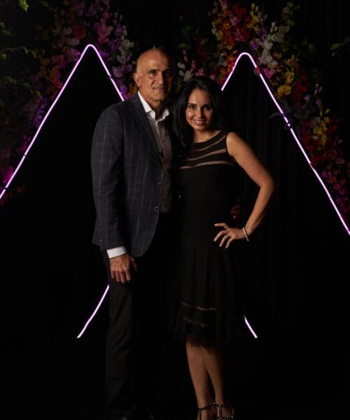 A man and woman are posing together in front of a dark backdrop adorned with neon pink lights in a triangular shape and floral decorations. The man is wearing a checked suit jacket and white shirt, while the woman is in a black dress with sheer segments.