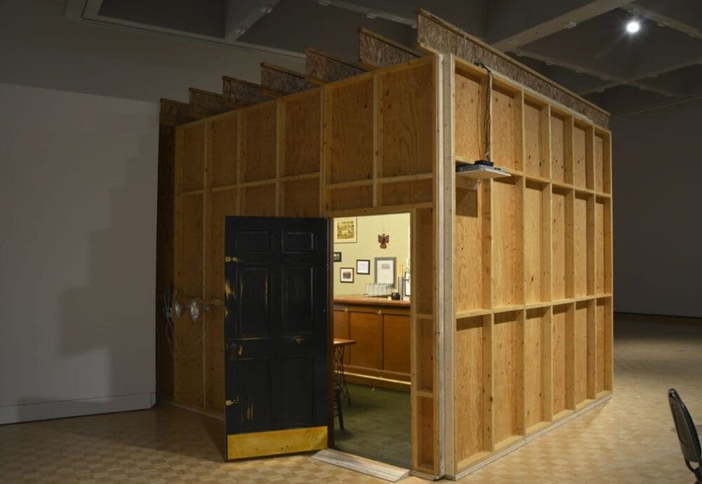A wooden structure resembling a small room or office, with an open dark-colored door revealing an interior with a desk, cabinets, and picture frames on the wall. The structure is located within a larger, well-lit space with checkered flooring.