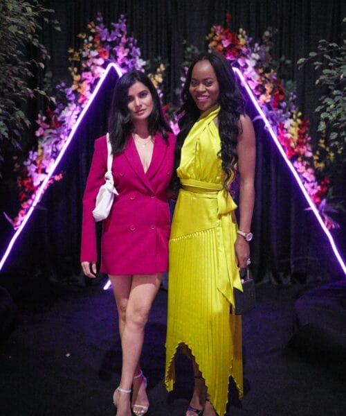 Two women posing in front of a decorative backdrop with colorful triangular neon lights. One wears a bright pink blazer dress and silver heels, holding a white handbag. The other wears a yellow pleated dress and gold heels, holding a black clutch.