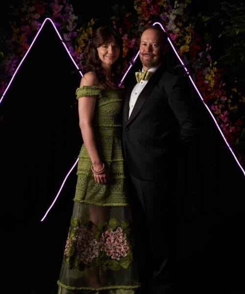 A woman in a green floral dress and a man in a black tuxedo with a bow tie stand close together, both smiling. They are in front of a dark backdrop with vibrant floral accents and neon pink triangular light decorations.