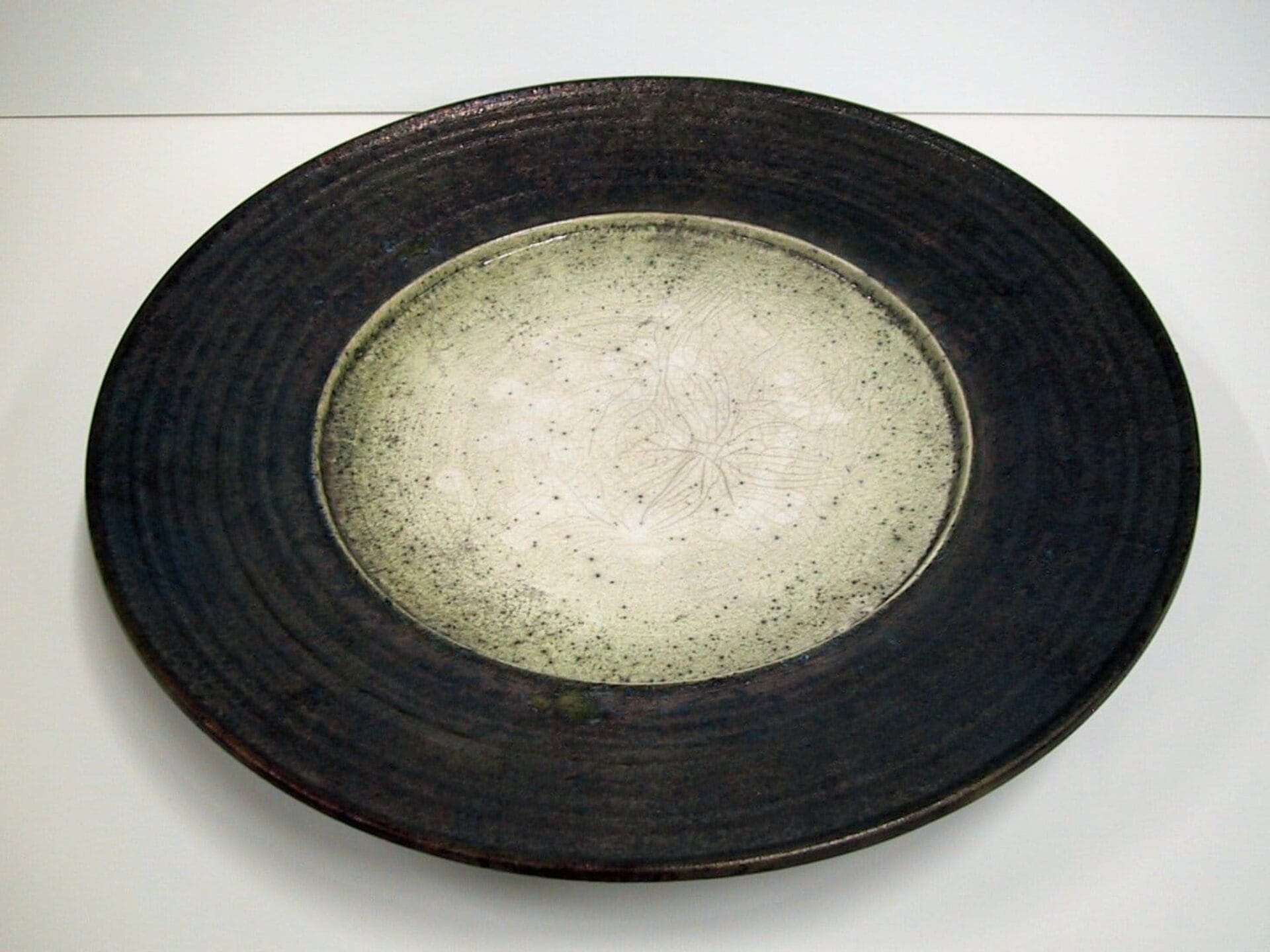 A wide, round ceramic bowl with a dark brown, textured rim and a lighter beige center featuring a delicate, abstract pattern. The bowl's design showcases a contrast between the rough exterior and the smooth, decorated interior.