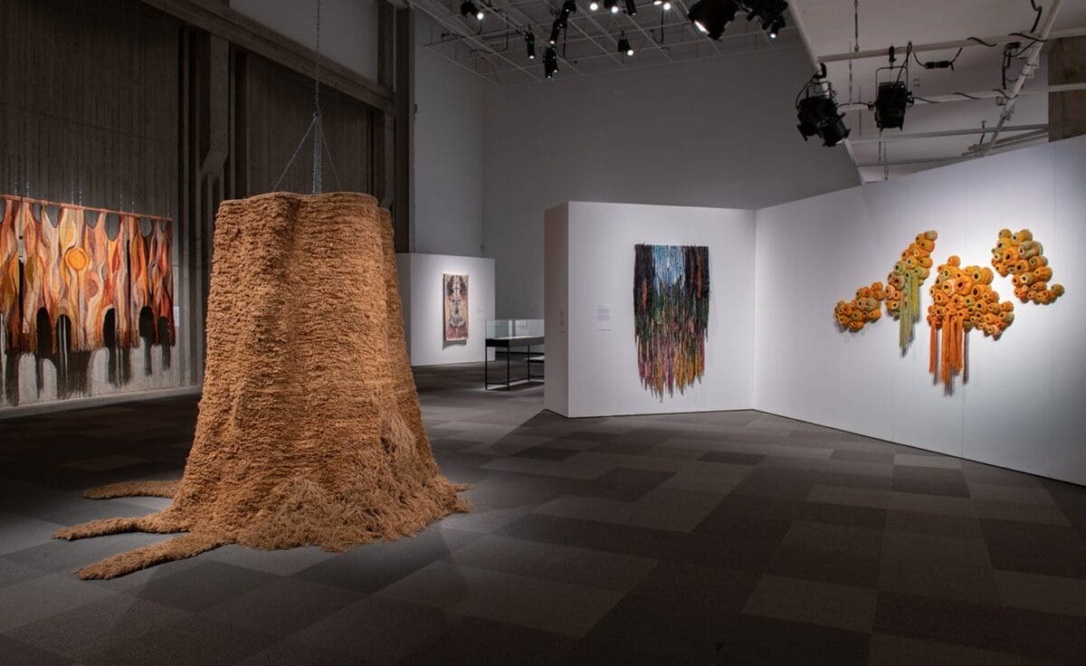 Large textile works on display, one looks like a brown tree stump, one on the wall looks like colourful barnacles, others can be seen in the background.