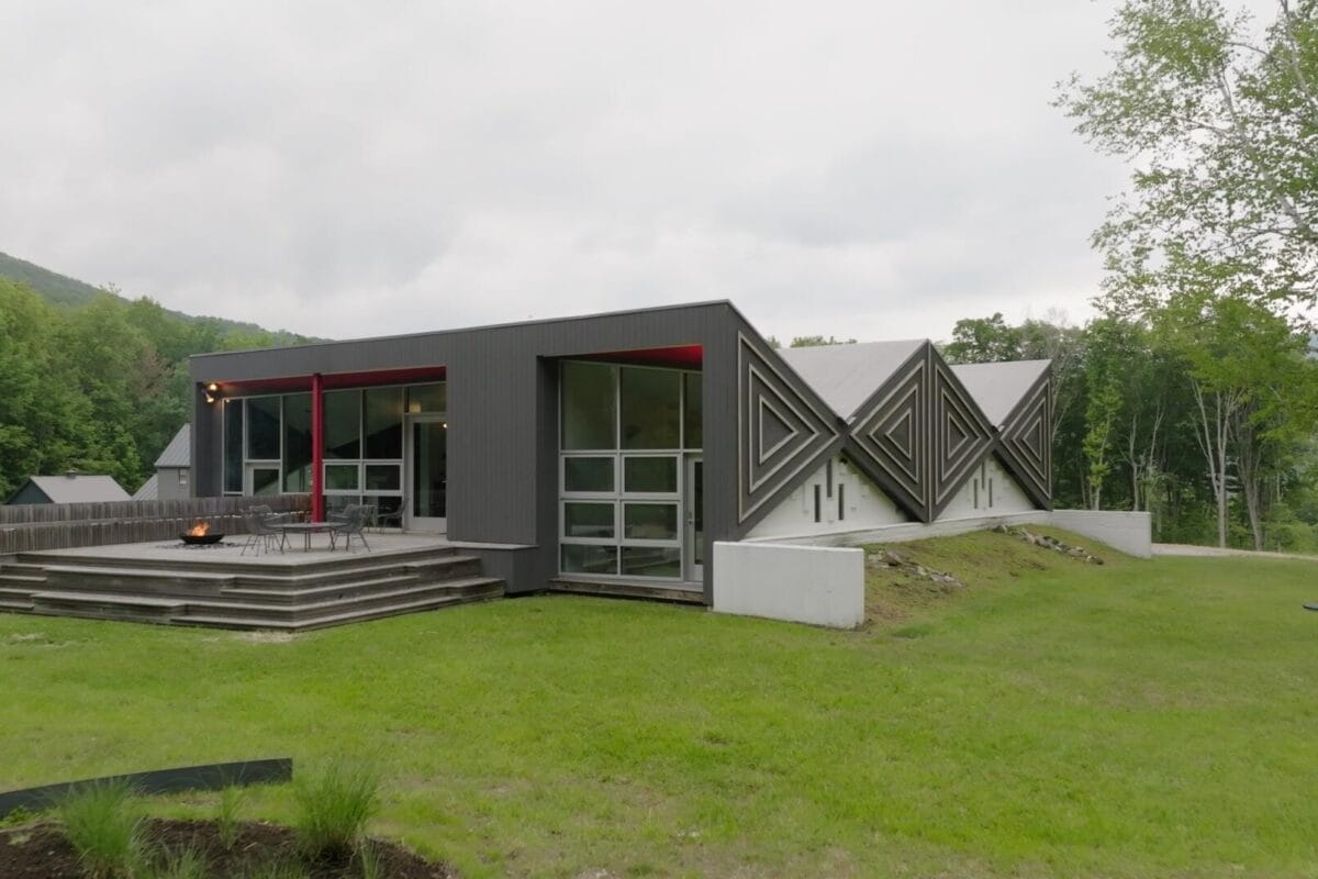 Une maison moderne et angulaire avec de grandes fenêtres et un design géométrique se trouve sur une pelouse herbeuse entourée d'arbres. L'extérieur présente des panneaux gris et des accents rouges, avec une terrasse en bois à l'avant. Le ciel est couvert.