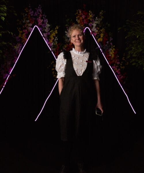 A person is standing in front of a dark backdrop with colorful, illuminated floral decorations and pink neon lights forming a mountain shape. They are wearing a black pinafore dress over a white, puffy-sleeved blouse and holding a phone in one hand, smiling.
