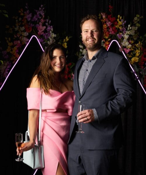 A woman in a pink dress and a man in a dark suit, both holding glasses of champagne, smile while standing in front of a floral background with neon triangular light decorations. They are surrounded by greenery and vibrant flowers.
