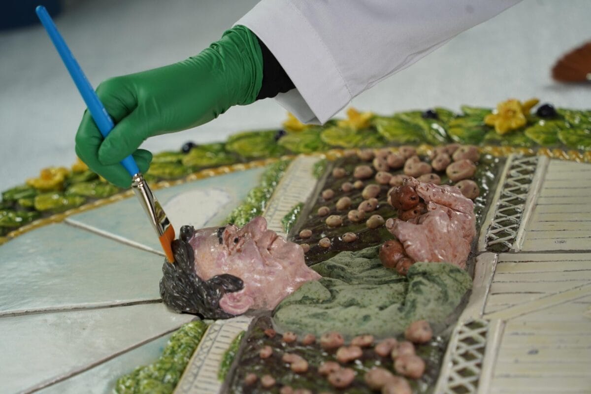A person wearing a green glove uses a paintbrush to touch up a ceramic artwork depicting a man surrounded by foliage and potatoes. The artwork features vibrant green and earthy tones with a circular floral border.