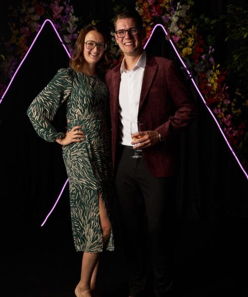 A smiling couple poses together against a dark background with neon lights and colorful flowers. The woman wears a green dress with beige patterns and beige heels, while the man wears a burgundy jacket over a white shirt and black pants, holding a glass of wine.
