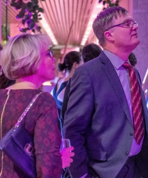 A group of people is attending an art exhibit. A man wearing glasses and a suit, along with a woman wearing a patterned dress and holding a drink, are closely examining a painting. The room is illuminated with purple lighting, and there are other artworks and attendees in the background.