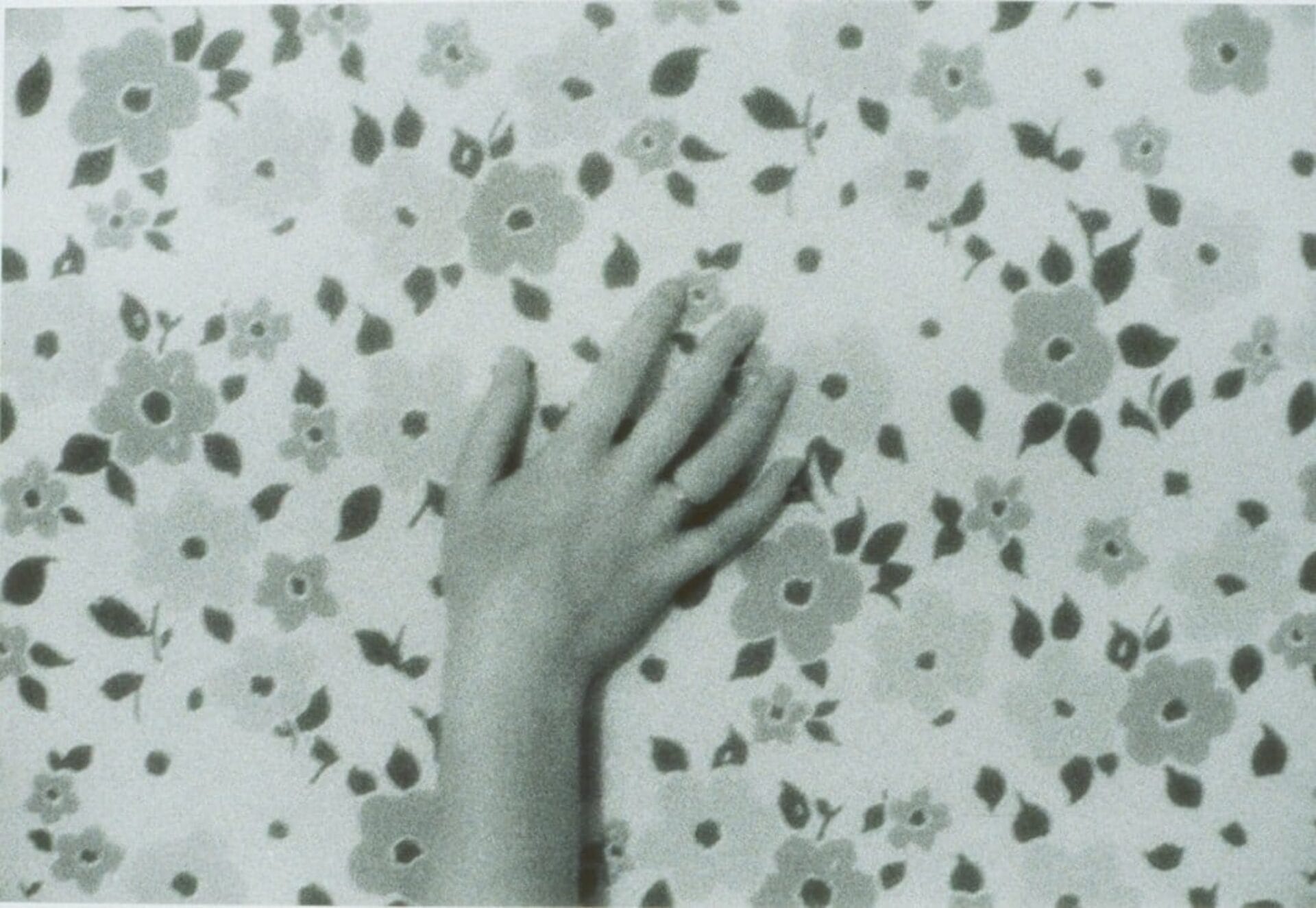 A grayscale image of a hand resting on a wall covered in a floral pattern. The hand is positioned with fingers slightly spread, blending softly with the background of flowers and leaves.