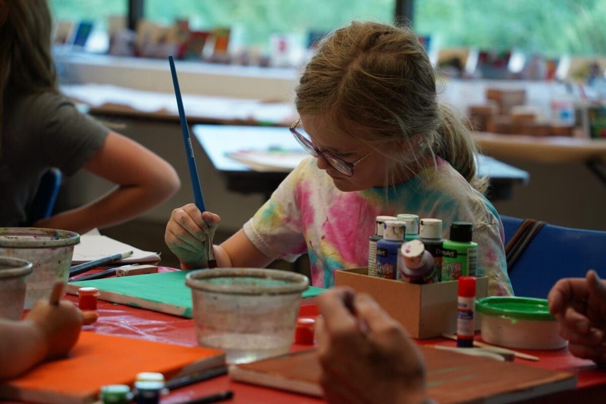 Une jeune fille portant des lunettes et une chemise tie-dye peint sur une toile verte à une table recouverte de fournitures d'art, de gobelets d'eau et de bouteilles de peinture dans une salle de classe bien éclairée.