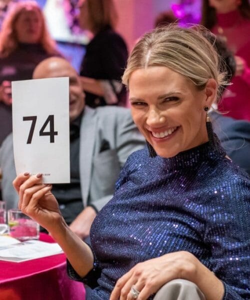 A woman with blonde hair smiles at the camera while holding up a card with the number 74. She is wearing a sparkly blue top and is sitting amongst a crowd in what appears to be a lively event or gathering. The background shows other people engaged and some holding drinks.