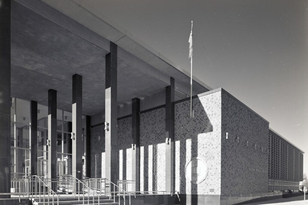 Black and white image of a modernist-style public building with large vertical pillars, a flat roof, and a brick exterior. The structure features a prominent entrance with a glass facade, multiple steps, and handrails. A flagpole is visible near the entrance, adding to the serene atmosphere reminiscent of Spring on the Prairies.