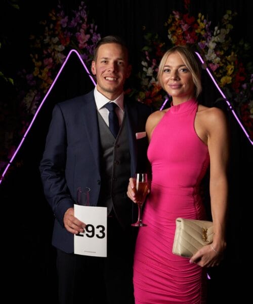 A man in a dark suit and tie holds an auction paddle number 93, standing next to a woman in a bright pink sleeveless dress holding a glass of wine and a beige clutch. They are posing in front of a dark backdrop with purple neon lights and floral decorations.