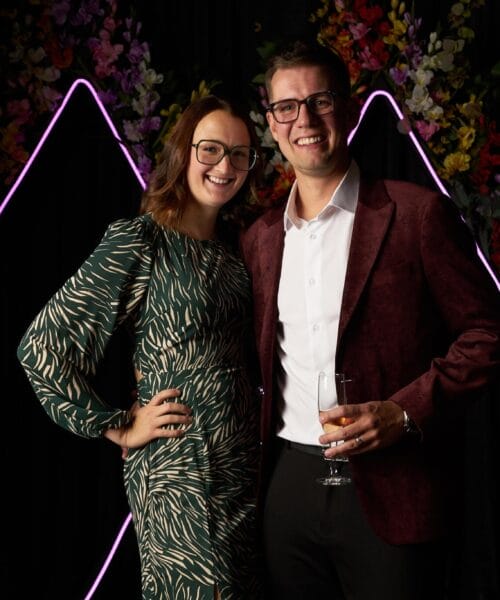 A smiling woman in a patterned green dress and a smiling man in a maroon jacket and white shirt stand together, holding a drink. They are posing in front of a neon-lit floral backdrop shaped like two triangles. The environment appears festive and elegant.