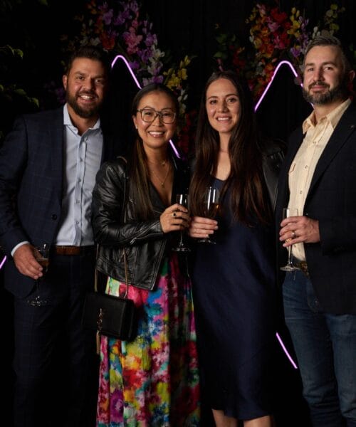 Four people dressed in semi-formal attire stand close together, smiling and holding champagne glasses. The background is decorated with neon purple lights and foliage.