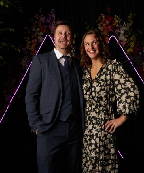 A man in a blue suit and tie stands next to a woman in a black and white floral dress. They are smiling and posing in front of a backdrop featuring neon purple triangles and colorful flowers, with lush green plants on either side.