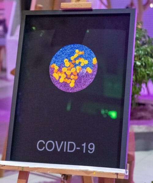An easel holds a poster depicting an artistic representation of the COVID-19 virus, featuring yellow particles on a blue and purple background, with 