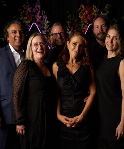 A group of six people, three men and three women, dressed in formal attire, are posing together and smiling. The background is decorated with green foliage, colorful flowers, and purple neon lights. The atmosphere appears festive and elegant.
