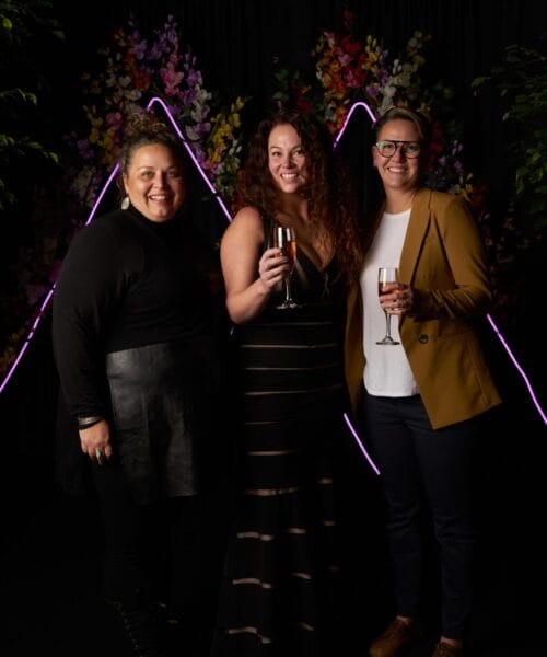 Three people are standing and smiling at a formal event. The person on the left is wearing a black outfit, the one in the middle is in a black dress and holding a drink, and the person on the right is in a tan blazer and glasses, also holding a drink. The background features colorful floral decorations and neon lights.