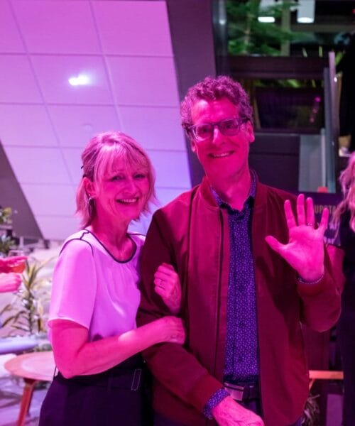 A smiling woman with blonde hair and a man with curly hair, wearing glasses and a red jacket, stand in a brightly lit room with purple lighting. The man waves at the camera while the woman holds his arm. They appear happy and are dressed casually.