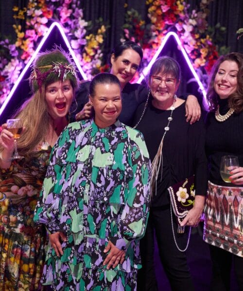 Five people pose for a photo in front of a backdrop decorated with vibrant flowers and illuminated purple triangles. They are all smiling and enjoying drinks, dressed in colorful and stylish outfits. Trees frame the sides of the image.