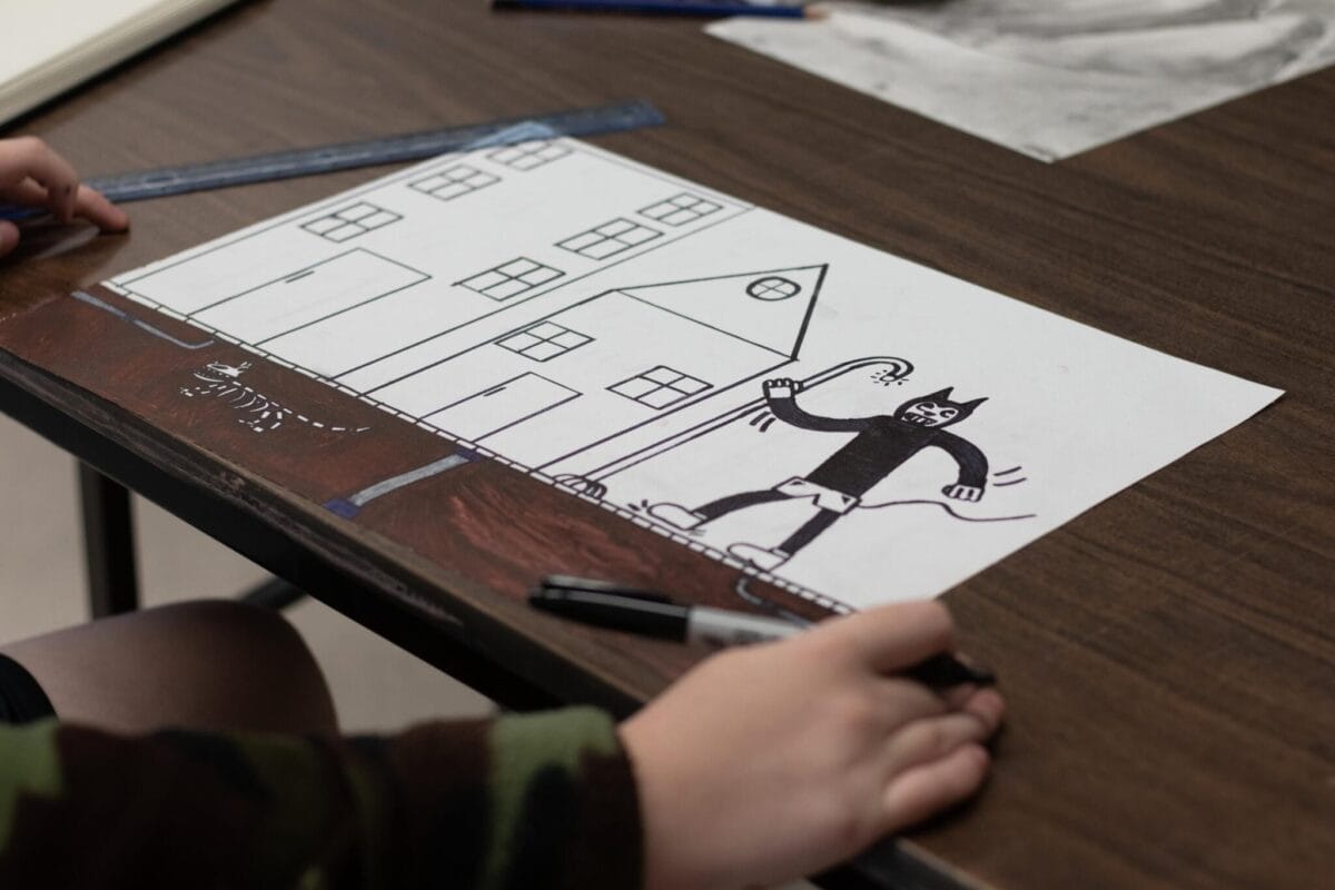 Une personne dessine un chat noir tenant une canne devant des maisons sur du papier blanc. Un squelette est également dessiné. Du matériel artistique et une règle sont posés sur la table marron.