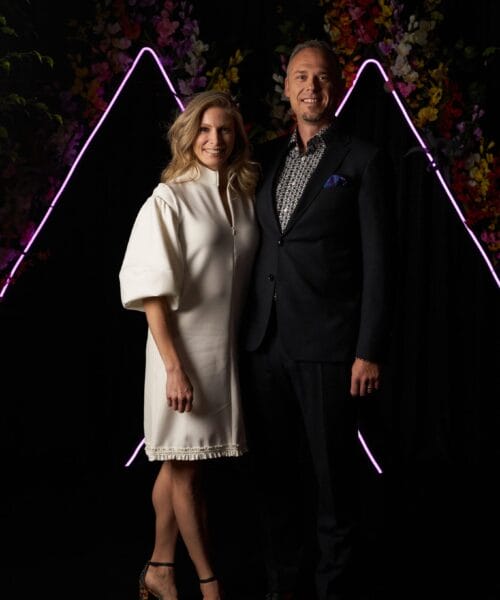 A woman in a white dress and a man in a dark suit stand together in front of a black backdrop with neon purple zigzag lines and colorful floral decorations. They are both smiling and facing the camera. The woman is wearing open-toed heels.