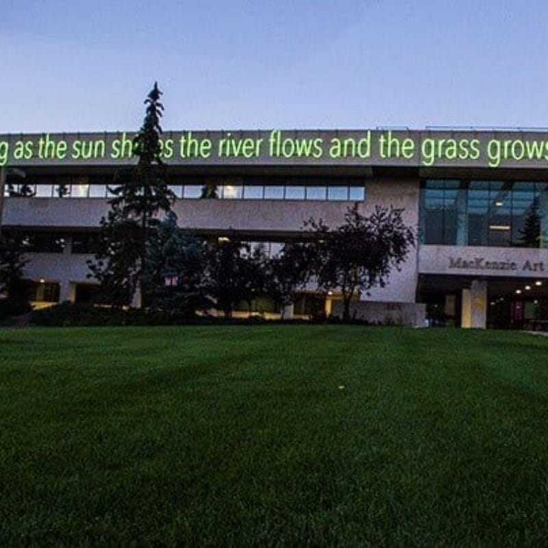 Un bâtiment avec un grand panneau électronique affichant le texte « tant que le soleil brille, la rivière coule et l'herbe pousse » près de son toit. Le bâtiment a de nombreuses fenêtres et il y a une zone herbeuse avec des arbres et une statue devant. L'entrée indique « MacKenzie Art Gallery.