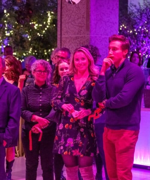 A group of people stands together in a dimly lit room with pink and purple lighting, watching something in front of them. Some hold drinks, and a few trees adorned with lights add to the atmosphere. Everyone appears engaged and focused on what they are observing.