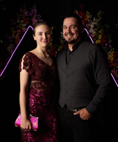 A woman in a red, lace dress stands next to a man in a gray vest and black shirt. They are smiling in front of a black backdrop with floral decorations and purple neon light accents. The woman holds a pink clutch. Both appear happy and well-dressed for an event.