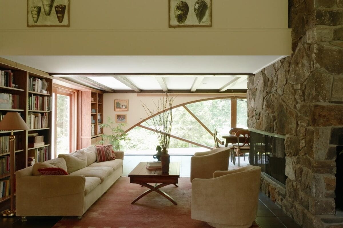Un salon chaleureux avec une grande cheminée en pierre, des canapés beiges et une table basse en bois sur un tapis rouge. Des étagères bordent un mur et de grandes fenêtres offrent une vue sur les arbres. La lumière du soleil illumine la pièce, créant une atmosphère chaleureuse.