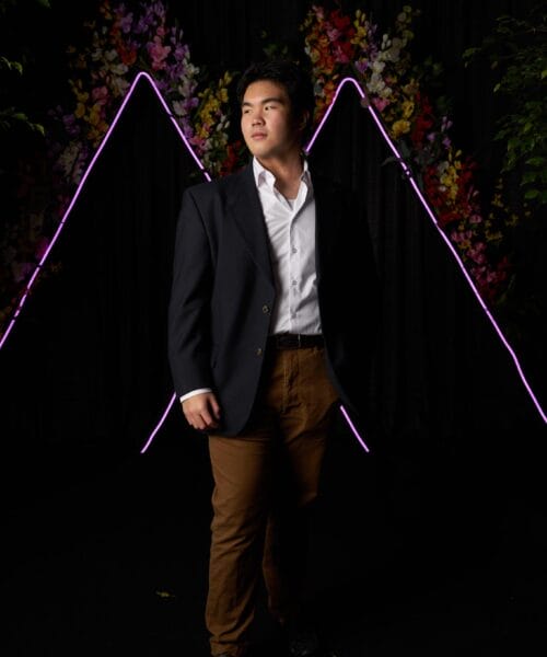 A person in a dark blazer, white shirt, and brown pants stands with one hand in his pocket. He is looking to his right. Behind him is a black backdrop adorned with colorful flowers and illuminated with V-shaped neon lights.