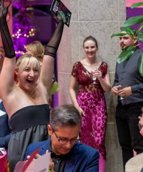 A joyful woman at a formal event raises her hands in celebration, holding a magazine. She is surrounded by other elegantly dressed people, some clapping, while others are engaged in conversation. The setting is adorned with vibrant pink lighting and plants.