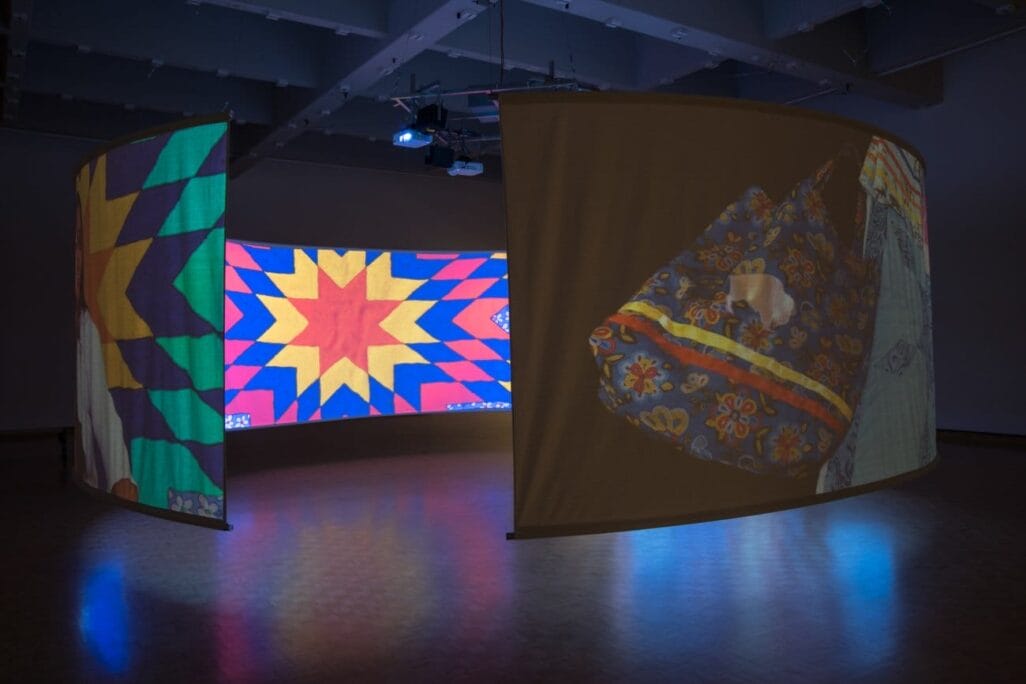 A dark room features a multimedia installation with three large curved screens projecting vibrant geometric patterns and colorful designs. The floor reflects the glowing patterns, enhancing the visual impact of the illuminated artwork.