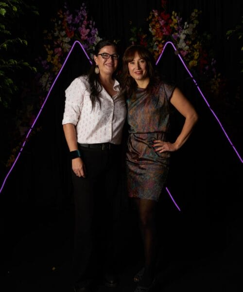 Two people are standing side by side in front of a dark backdrop with neon purple triangular shapes and colorful flowers. One is wearing a white shirt and black pants, while the other is in a colorful dress. Both are smiling and posing for the camera.