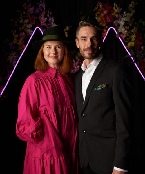 A woman in a bright pink dress and green hat stands next to a man in a black suit with a white shirt and pocket square. They are posing against a black backdrop adorned with neon pink triangular lights and colorful flowers.