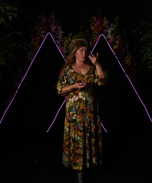 A woman stands against a dark backdrop with vibrant purple neon triangles and colorful flowers. She wears a long, floral dress and a headband, holding a small object with one hand and gesturing near her face with the other. Plants frame the sides of the scene.