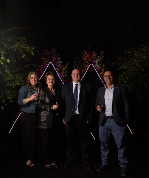 Four people are standing and posing for a photograph in front of a backdrop with pink illuminated triangular shapes. They are dressed in semi-formal attire and smiling while holding drinks. Green foliage is visible on both sides of the group.